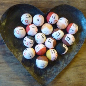 May include: A collection of small, round, hand-painted beads with closed eyes and various hair colors, resting in a heart-shaped metal dish. The beads have red, black, brown, and yellow accents. The dish has a rustic, aged appearance.