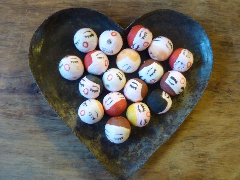 May include: A collection of small, round, hand-painted beads with closed eyes and various hair colors, resting in a heart-shaped metal dish. The beads have red, black, brown, and yellow accents. The dish has a rustic, aged appearance.
