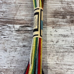 May include: Bundle of 32 pipe cleaners in various colors, including red, yellow, green, and blue. The pipe cleaners are bundled together with a paper label that has text on it. The background is a weathered, white-washed wooden surface.