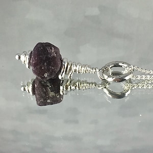 Puede incluir: Un collar de cadena de plata con un colgante de piedra preciosa de color rojo oscuro. La piedra preciosa está envuelta en alambre de plata y tiene una textura rugosa y natural.