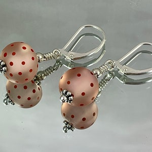 Puede incluir: Un par de pendientes de plata con una cuenta de vidrio rosa y roja con lunares en cada pendiente.