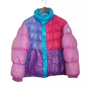 Puede incluir: Una chaqueta acolchada multicolor con un esquema de color rosa, azul y morado. La chaqueta tiene una cremallera y un cierre de botón a presión. La chaqueta tiene un parche de logotipo en la manga izquierda.