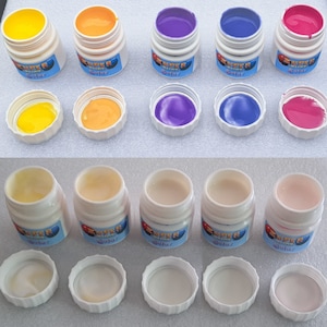 Könnte beinhalten: Eine Sammlung kleiner, weißer Gläser mit farbigen Farben. Die Farben umfassen Gelb, Orange, Lila, Blau und Rosa. Die Gläser sind mit "SUPER Solar" beschriftet. Einige Deckel sind abgenommen und zeigen die Farbe. Maße nicht zutreffend.