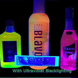 Könnte beinhalten: Drei Glasflaschen mit Likör, eine gelbe, eine rosa und eine orange, werden unter Schwarzlicht beleuchtet. Die Flaschen sind mit "Blavod", "Canti" und "Super" beschriftet.