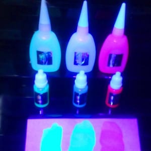 Peut inclure: Trois flacons de colle qui brillent dans le noir, en bleu, blanc et rose. Les flacons sont disposés en rangée, le flacon bleu à gauche, le flacon blanc au milieu et le flacon rose à droite. Il y a trois flacons plus petits de peinture qui brillent dans le noir, en bleu, vert et rouge. La peinture est présentée sur une surface, qui brille sous une lumière noire.