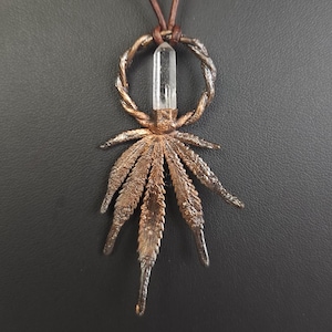 May include: A bronze pendant necklace with a cannabis leaf design. The pendant features a clear crystal and a twisted metal ring. The necklace has a brown leather cord.
