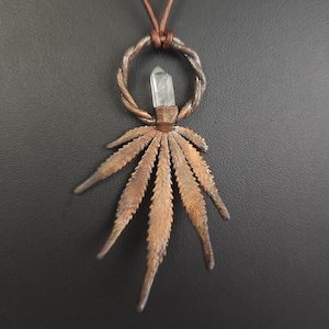 May include: A copper pendant necklace with a cannabis leaf design. The pendant features a clear crystal and a twisted metal ring. The necklace hangs from a brown cord. The background is dark grey.