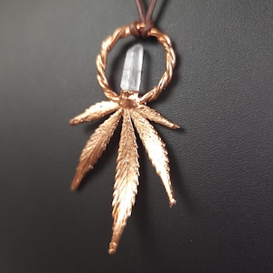 May include: A copper-toned pendant necklace with a cannabis leaf design. The pendant incorporates a clear crystal and a twisted ring. The necklace hangs from a brown cord. The background is a dark, solid colour.