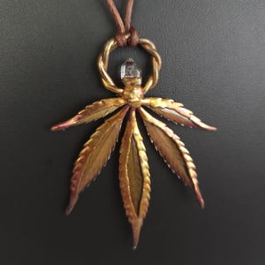 Puede incluir: Collar con colgante artesanal con diseño de hoja de cannabis color bronce. El colgante tiene un cristal transparente en el centro y está suspendido de un cordón de cuero marrón. El collar está sobre un fondo oscuro.