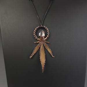 Könnte beinhalten: Kupferfarbene Anhängerkette mit Cannabisblatt-Design. Der Anhänger hat einen klaren Kristall an der Spitze und hängt an einer schwarzen Kordel. Das Blatt hat detaillierte Adern und eine strukturierte Oberfläche.