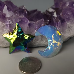 May include: Two iridescent glass pieces, a star and a crescent moon, on a purple amethyst crystal cluster. The star is green, blue, and purple. The moon is blue and iridescent.