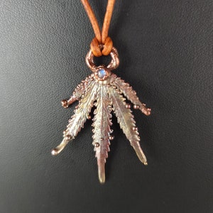 Op de afbeelding: Een koperkleurige ketting met een gedetailleerd bladontwerp en een kleine iriserende edelsteen in het midden. De ketting is aan een bruin leren koord.