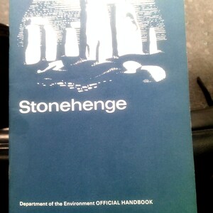 Puede incluir: Un folleto azul con la palabra "Stonehenge" en blanco. La portada presenta una ilustraci&oacute;n blanca del monumento de Stonehenge bajo un arco blanco. El texto "Department of the Environment OFFICIAL HANDBOOK" est&aacute; debajo.