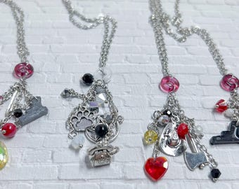 May include: Four silver necklaces with decorative charms. Each necklace has a chain with a pink bead at the top, and a collection of charms including a heart, axe, and other shapes. The charms are accented with red, black, and clear beads.