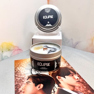 May include: A silver candle tin with a black label that reads "Serpent & Flame" and "Eclipse - Water Grapefruit -"  The candle is lit and has a white and black wax with gold glitter. The candle is sitting on a white box and a white stand.