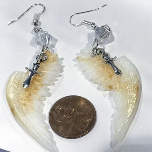 Puede incluir: Un par de pendientes colgantes con un dise&ntilde;o de ala de &aacute;ngel blanco y dorado, un dije de daga plateada y una cuenta de cristal. Los pendientes son aproximadamente del tama&ntilde;o de una moneda de un centavo de euro.