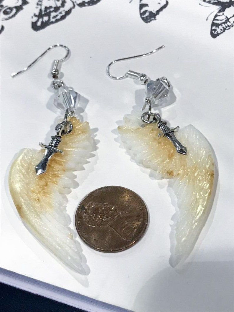 Puede incluir: Un par de pendientes colgantes con un dise&ntilde;o de ala de &aacute;ngel blanco y dorado, un dije de daga plateada y una cuenta de cristal. Los pendientes son aproximadamente del tama&ntilde;o de una moneda de un centavo de euro.