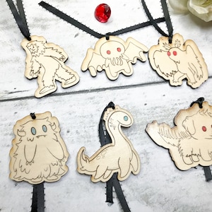 May include: Six wooden ornaments with black ribbon hangers. Each ornament features a different mythical creature: a yeti, a Cthulhu-like monster, a bird-like creature, a furry monster, a plesiosaur, and a dog.