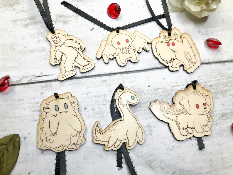 May include: Six wooden ornaments with black ribbon hangers. Each ornament features a different mythical creature: a yeti, a Cthulhu-like monster, a bird-like creature, a furry monster, a plesiosaur, and a dog.