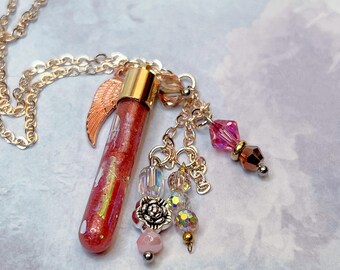 Könnte beinhalten: Goldfarbene Halskette mit Anhänger. Der Anhänger ist ein kleines, rot glitzerndes Fläschchen mit goldenem Verschluss und kupferfarbenem Flügel-Charm. Zusätzliche Charms sind rosa und irisierende Perlen sowie eine silberne Rose.