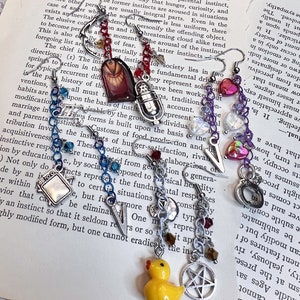 Pode incluir: Uma coleção de brincos artesanais com vários pingentes, incluindo um patinho de borracha amarelo, um pentagrama e um coração. Os brincos apresentam correntes e contas prateadas, criando um estilo único e eclético.