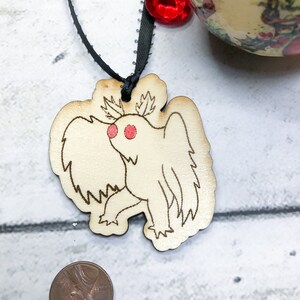 May include: A wooden ornament shaped like a moth with red eyes. The ornament is hanging from a black ribbon with a red jewel.