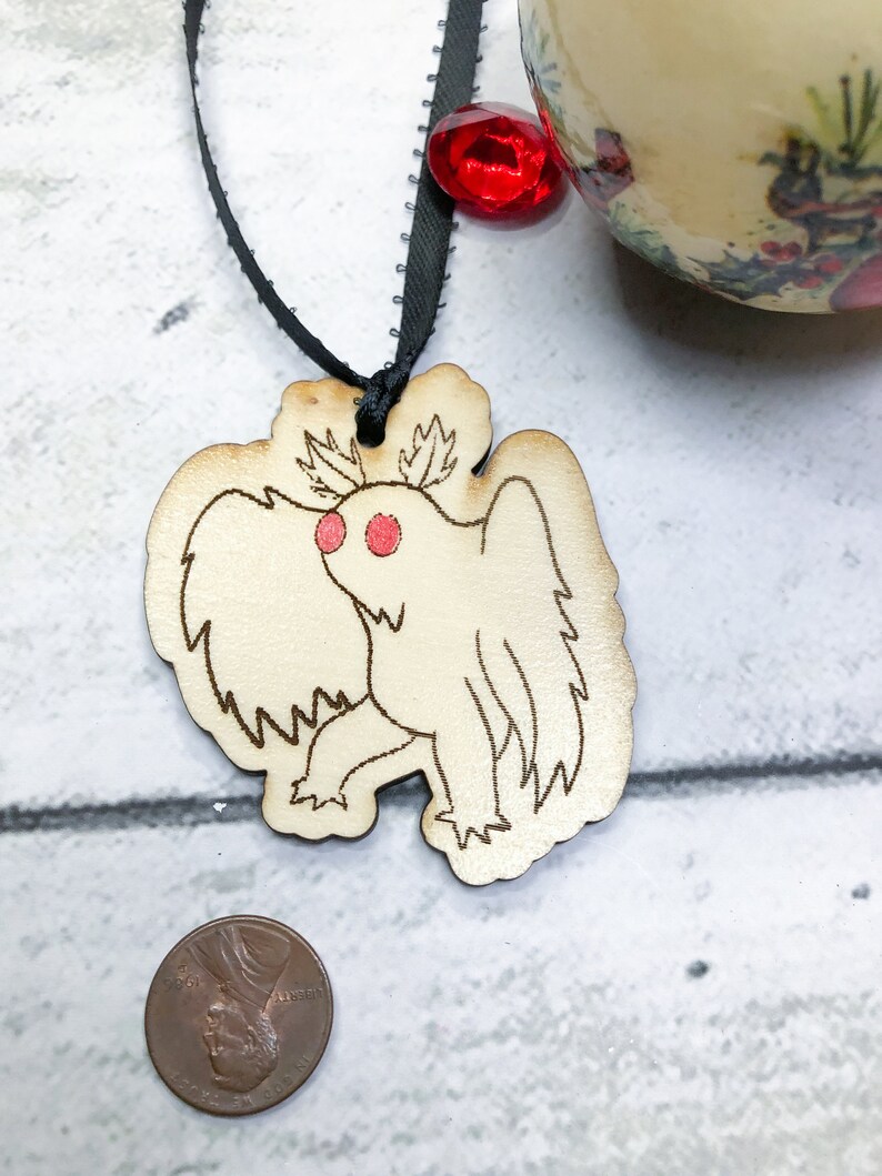 May include: A wooden ornament shaped like a moth with red eyes. The ornament is hanging from a black ribbon with a red jewel.