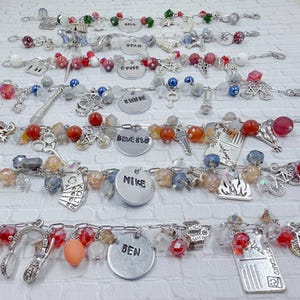 Puede incluir: Varios brazaletes con dijes con varias cuentas y dijes. Los brazaletes presentan cadenas plateadas, cuentas de colores y dijes como bicicletas, llaves y etiquetas con nombres. Las etiquetas con nombres tienen nombres como "Ryan", "Evie", "Mike" y "Ben".