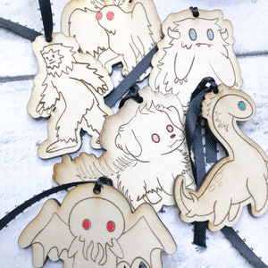 May include: Six wooden ornaments with black ribbon hangers. Each ornament features a different cartoon monster, including a yeti, a dog, a Cthulhu-like creature, a winged creature, a dinosaur, and a fuzzy monster.