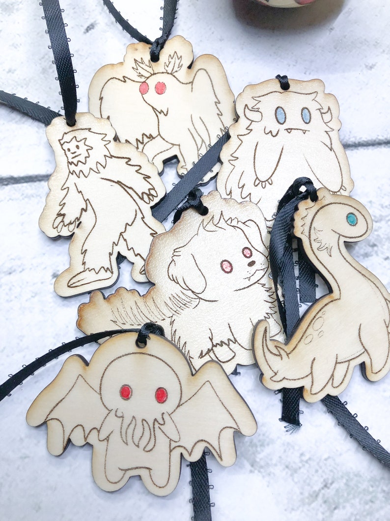 May include: Six wooden ornaments with black ribbon hangers. Each ornament features a different cartoon monster, including a yeti, a dog, a Cthulhu-like creature, a winged creature, a dinosaur, and a fuzzy monster.