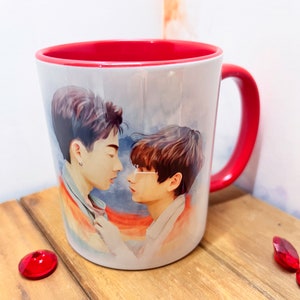 May include: A white ceramic mug with a red rim and handle. The mug features a watercolor illustration of two men embracing, with a rainbow flag in the background.