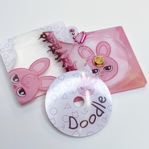 May include: A pink and white acrylic charm featuring a bunny design and the word "Doodle" inscribed on it. The charm has a hole in the centre and is attached to a pink and silver chain.