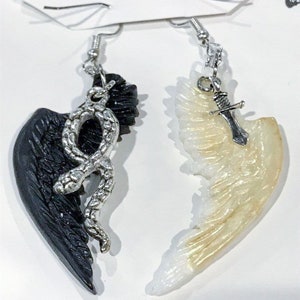 Puede incluir: Un par de pendientes con un ala negra con una serpiente plateada y un ala blanca con un dije de espada plateada.