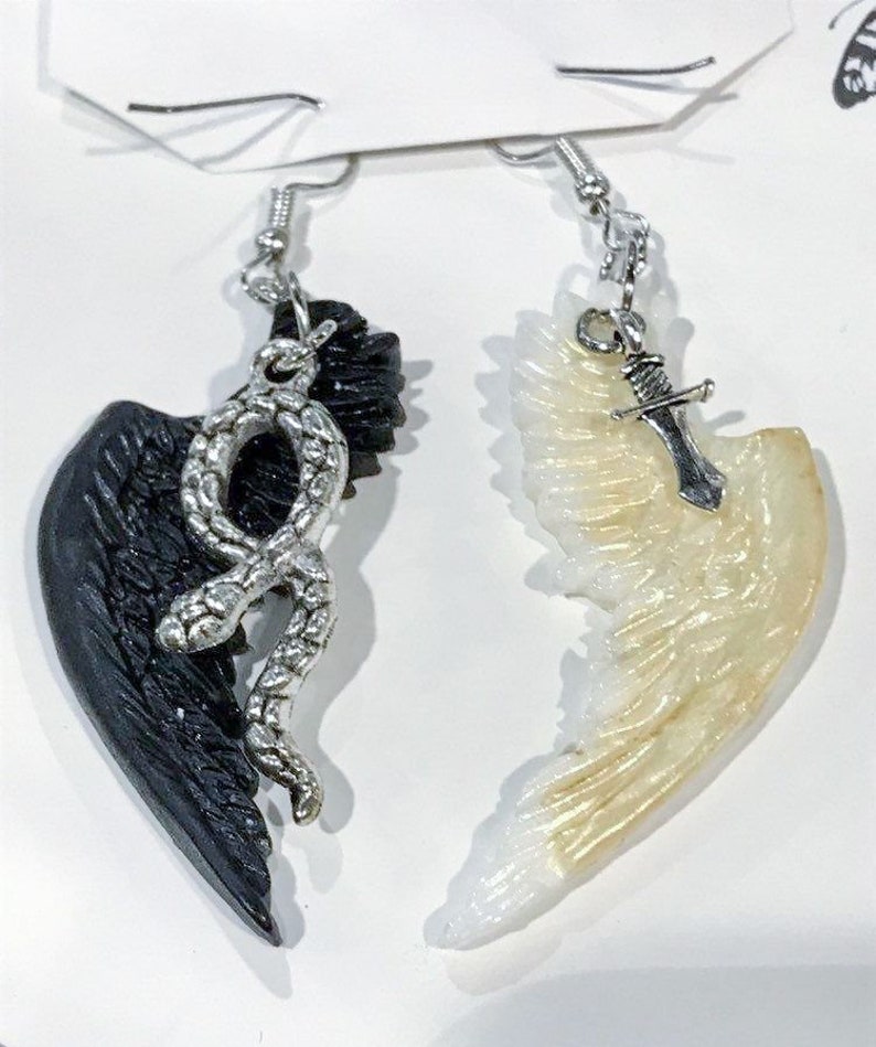 Puede incluir: Un par de pendientes con un ala negra con una serpiente plateada y un ala blanca con un dije de espada plateada.