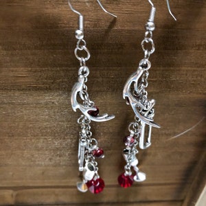 May include: Silver dangle earrings with red crystal accents. The earrings feature a crescent moon design with a chain and charms.