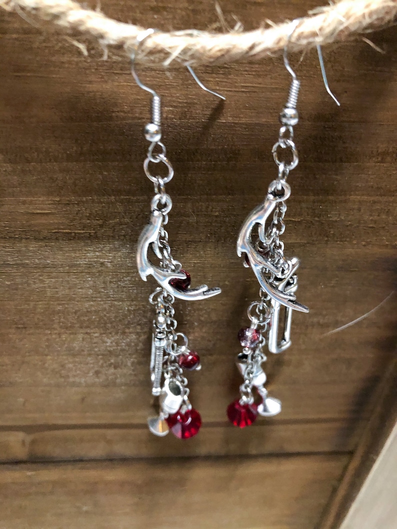 May include: Silver dangle earrings with red crystal accents. The earrings feature a crescent moon design with a chain and charms.