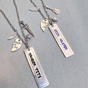 Puede incluir: Tres collares de plata con dijes. Los dijes incluyen un corazón roto con las palabras "Part of Cr", una guitarra, una espada, un pequeño ala de ángel, un corazón con las palabras "Ners in Time", y dos etiquetas rectangulares con las palabras "Danger Tits" y "Dick Master".