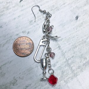 May include: Silver charm earring with a variety of charms, including a saw, a goblet, a crescent moon, and red and pink beads. The earring is about the size of a US penny.