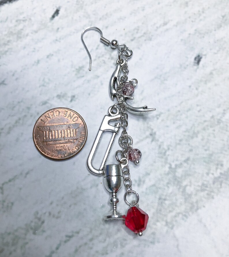 May include: Silver charm earring with a variety of charms, including a saw, a goblet, a crescent moon, and red and pink beads. The earring is about the size of a US penny.