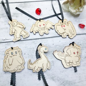 May include: Six wooden ornaments featuring different mythical creatures, including a yeti, a Cthulhu-like monster, a griffin, a yeti, a plesiosaur, and a dog with wings. Each ornament is attached to a black ribbon.
