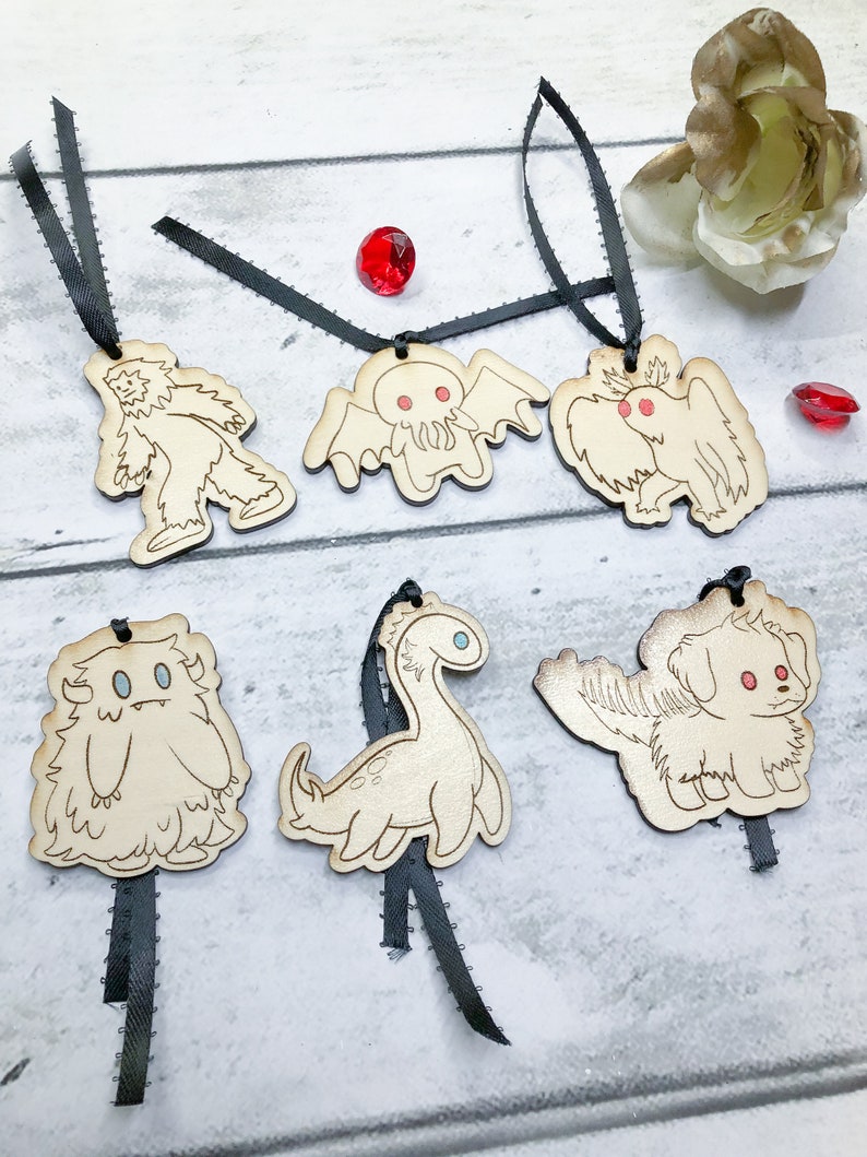 May include: Six wooden ornaments featuring different mythical creatures, including a yeti, a Cthulhu-like monster, a griffin, a yeti, a plesiosaur, and a dog with wings. Each ornament is attached to a black ribbon.