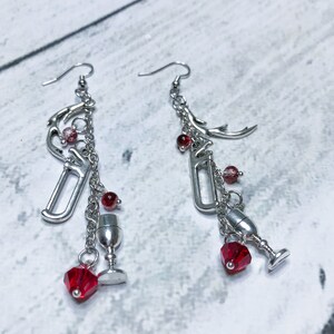 May include: A pair of silver dangle earrings with red crystal beads and charms, including a wine bottle, wine glass, and antlers.