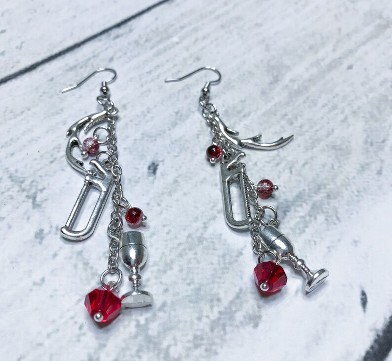 May include: A pair of silver dangle earrings with red crystal beads and charms, including a wine bottle, wine glass, and antlers.