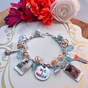Op de afbeelding: Een zilveren armband met verschillende bedels, waaronder een microfoon, een lichtstok, een foto van twee mensen en een bedel met de tekst "BTS" in rode en blauwe letters. De armband heeft ook verschillende heldere en blauwe glazen kralen.