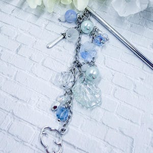 May include: A silver hair stick with a dangling charm featuring blue and clear beads, faux pearls, and a cloud-shaped charm. The hair accessory has a decorative floral design and is set against a white brick background.