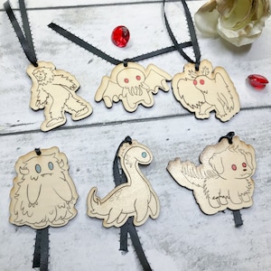 May include: Six wooden ornaments with black ribbon hangers. The ornaments feature various mythical creatures, including a yeti, a Cthulhu-like creature, a griffin, a sasquatch, a plesiosaur, and a dog.