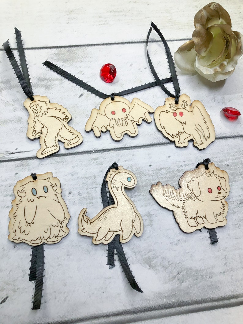 May include: Six wooden ornaments with black ribbon hangers. The ornaments feature various mythical creatures, including a yeti, a Cthulhu-like creature, a griffin, a sasquatch, a plesiosaur, and a dog.
