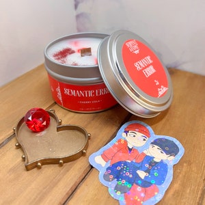 May include: A red and silver candle tin with the text "Semantic Error" and "Cherry Cola" on it. The candle is lit and has a red wax pool. There is a red gem in a heart-shaped frame and a sticker with two cartoon characters on a wooden surface.