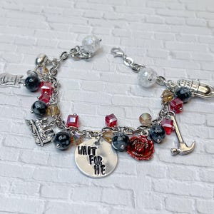 Puede incluir: Una pulsera de plata con varios dijes, incluyendo una guitarra, un micrófono y un martillo. La pulsera también presenta cuentas de cristal rojas y transparentes, cuentas negras y un dije redondo con el texto "WAIT FOR ME".