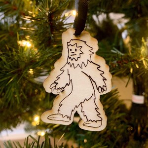 May include: A wooden Christmas ornament shaped like a Bigfoot, with a black ribbon attached. The ornament is hand-drawn in black ink and features a simple design of a Bigfoot walking.